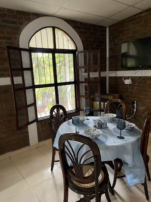 A dining area with a brick wall, a large arched window, a TV mounted on the wall, and a table set with bowls and glasses.