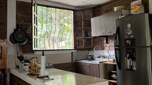 A kitchen with a brick accent wall, a stainless steel refrigerator, and a window with a view of greenery. - A place to enjoy nature 1-bedroom cabin in soothing Los Planes De Renderos. (Panchimalco)