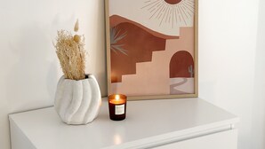 A white dresser with a framed artwork depicting a stylized sun and landscape, a vase with dried plants, and a lit candle.