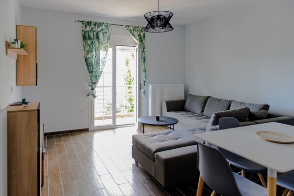 A modern living room with a sofa, dining table, and chairs, a wooden cabinet, and a large window with curtains.