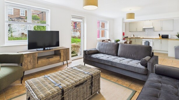 Ein modernes Wohnzimmer mit Fernseher, grauem Sofa, Rattan-Pouff und einer Küche im Hintergrund.