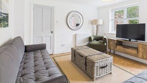 Ein modernes Wohnzimmer mit einem grauen Sofa, einem Korbsessel, einem Fernseher auf einem Holzboden, einer Lampe und einem Fenster mit Jalousien.