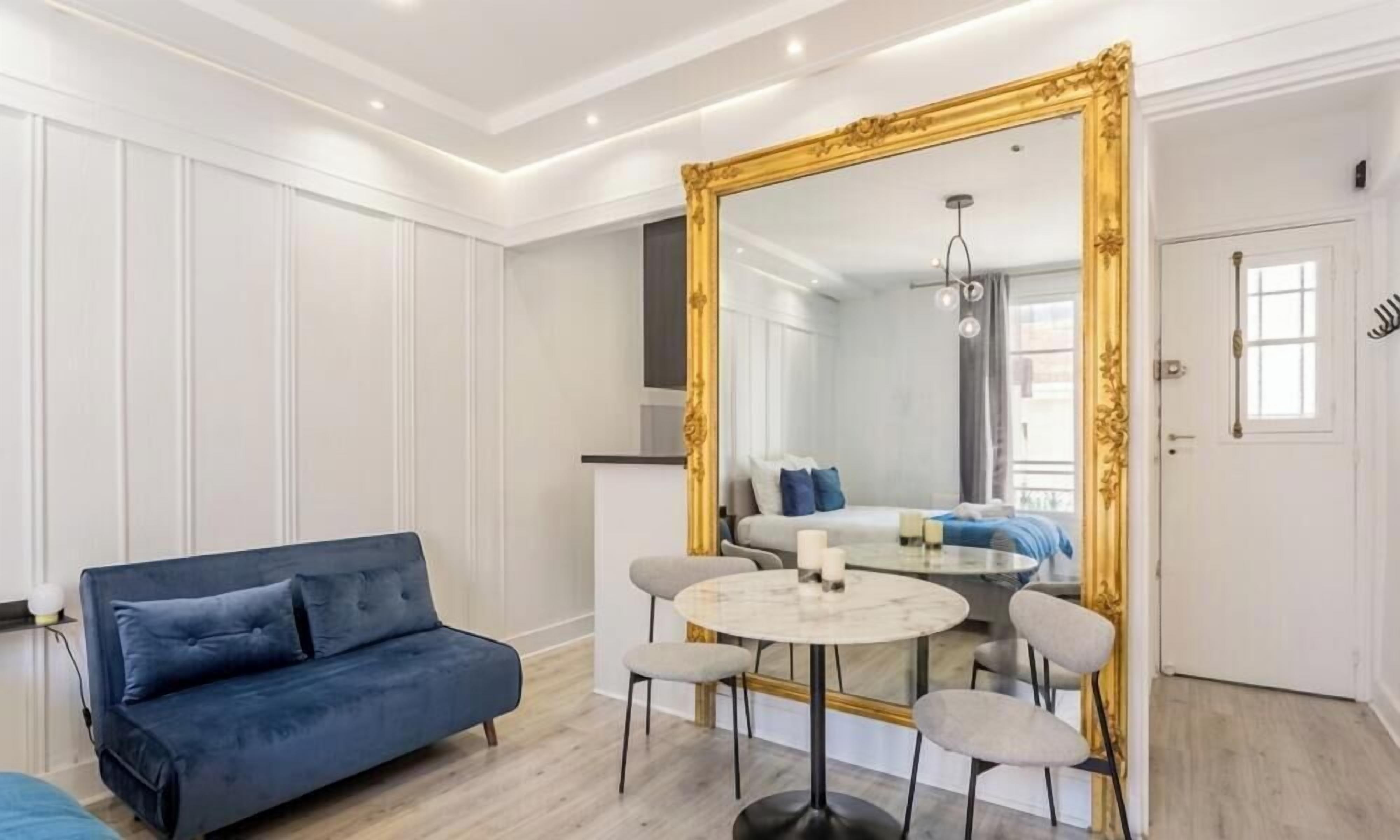 Una habitación moderna con un sofá azul, una mesa redonda con sillas, un espejo grande y una puerta con ventanal.