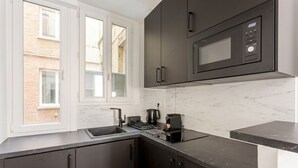 A modern kitchen with dark cabinets, a microwave, a coffee maker, and a window with a view of a building outside. - celine 18 (Paris)