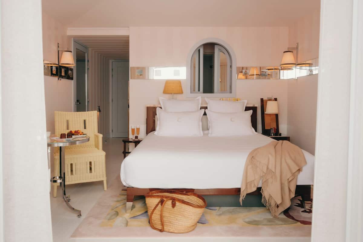 Un dormitorio ordenado con cama, mesitas de noche, una silla y una pequeña mesa con una cesta.