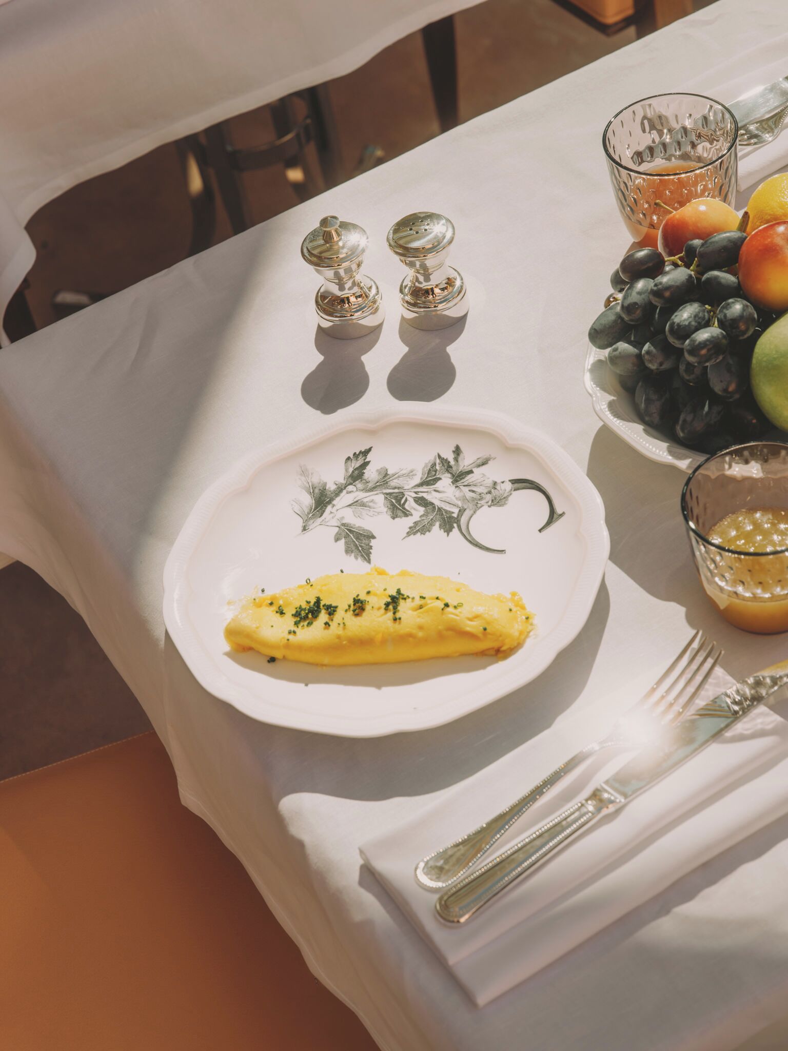 Una mesa dispuesta con un plato de comida, un bol de fruta, saleros y pimenteros, un vaso de jugo y cubiertos.