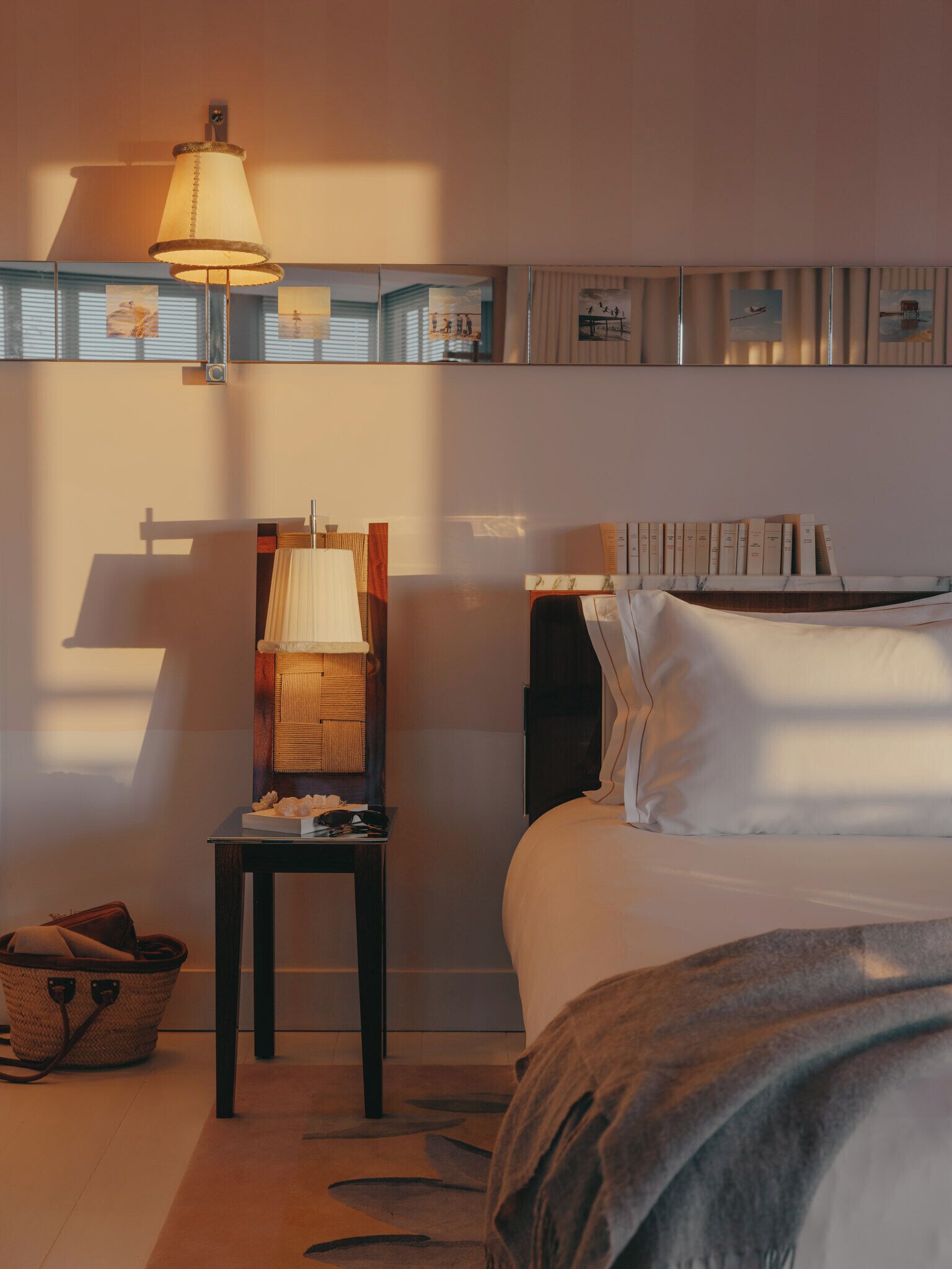 Une chambre d’hôtel avec un lit, une table de chevet avec une lampe, une étagère fixée au mur avec des livres et un panier posé sur le sol.