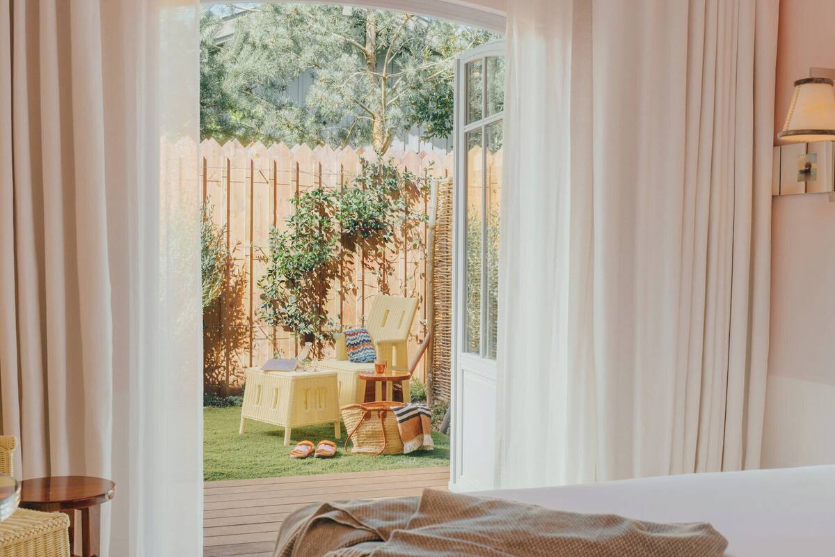 Un dormitorio con un ventanal grande, una cama, una silla de mimbre, una mesita y vistas a un jardín cercado.