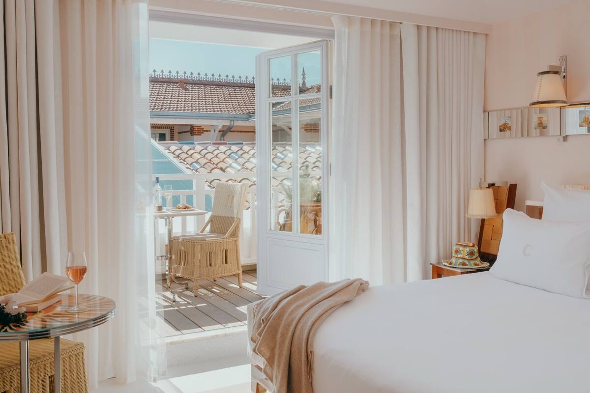 Habitación de hotel con cama, una mesa de vidrio con una silla, una mesa pequeña con una silla y vistas a un edificio con tejado de tejas.