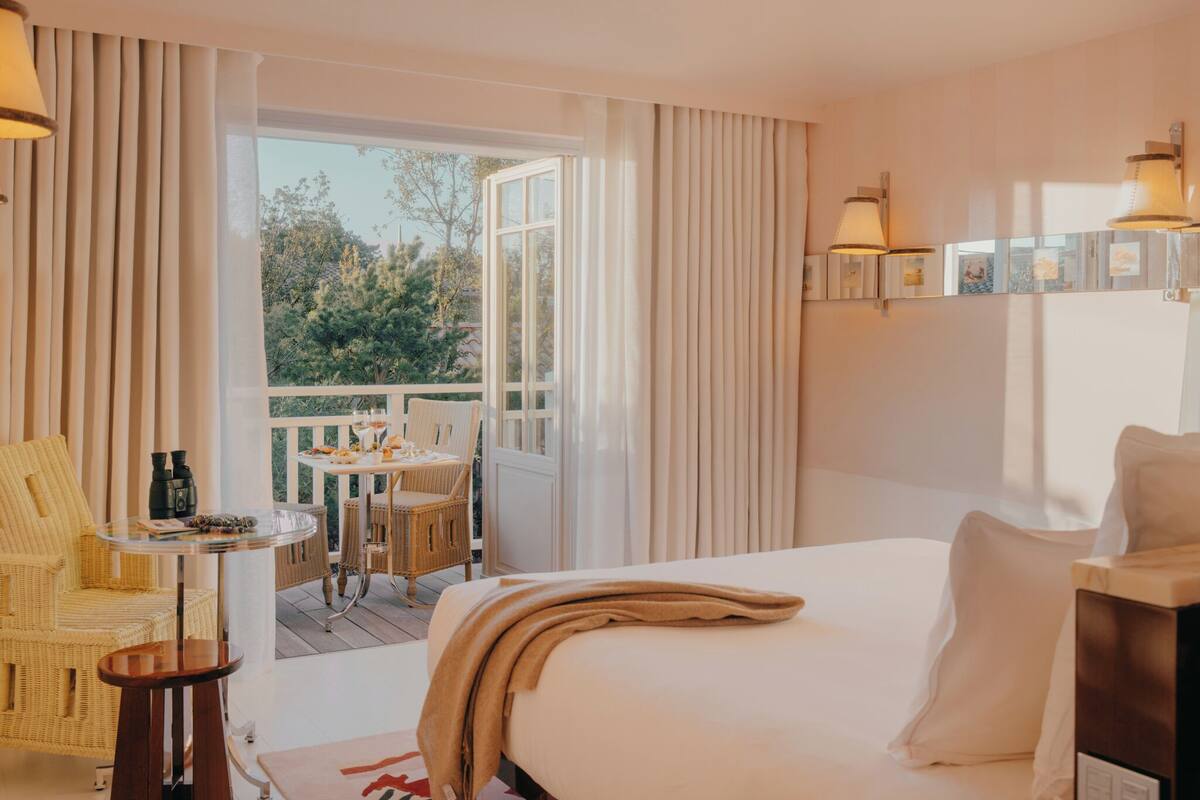 Habitación de hotel con cama, una pequeña mesa con sillas, un balcón con cortinas y vistas a árboles.