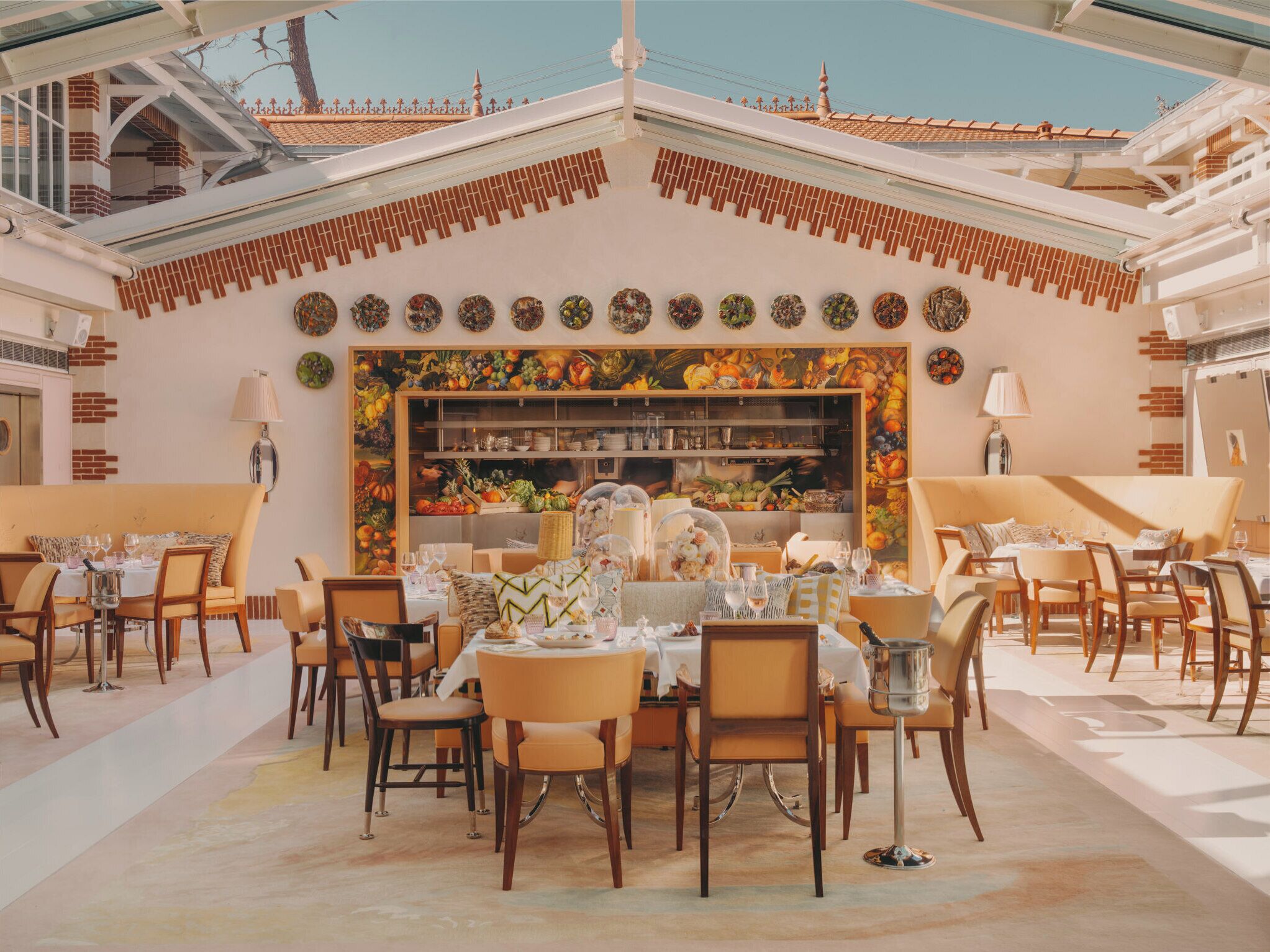 A dining area with a central table surrounded by chairs, a kitchen area with a fruit display, and a view of the sky through a glass ceiling.