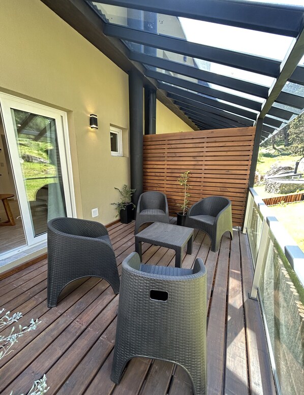 A covered outdoor seating area with wicker furniture, a wooden deck, and a view of a natural landscape.
