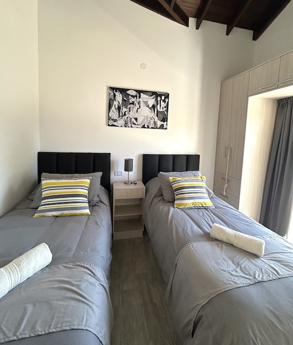 A room with two single beds, a nightstand with a lamp, a wall-mounted artwork, and a wardrobe.