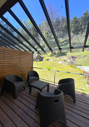 A covered outdoor seating area with wicker chairs and a table, overlooking a grassy landscape with trees and a rock formation.