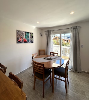 A dining area with a round table, wooden chairs, a painting on the wall, and a view through a window.