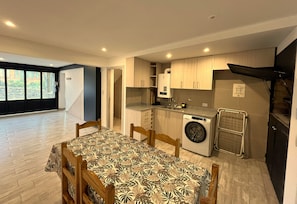 A compact kitchen with a washing machine, a dining table with chairs, and a view of a garden through a window.