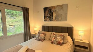 A hotel room with a large bed, two bedside tables with lamps, a window with curtains, and a painting on the wall. - Enjoy with your family the best view of the waterfall I Ayres del Bosque Accommodations (San Martín de los Andes)
