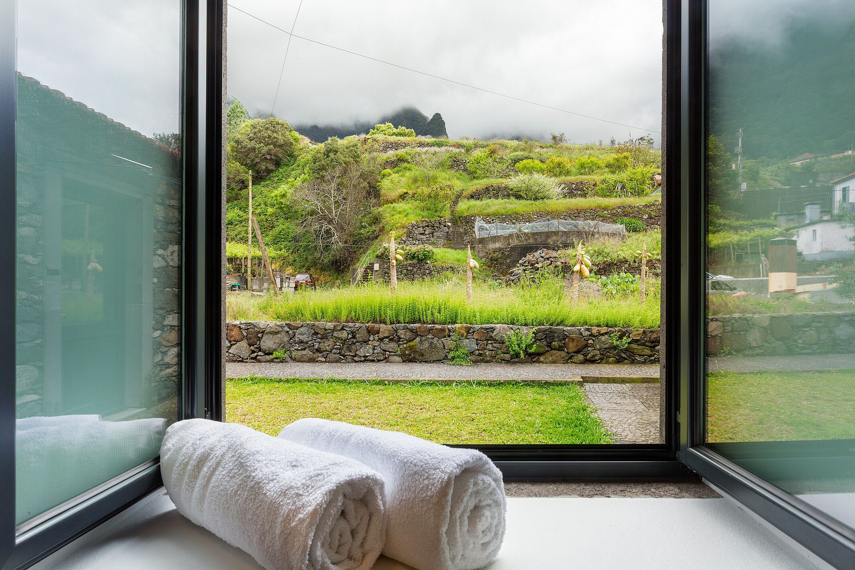 A room with a large window, a rolled white towel on the windowsill, and a stone wall outside.