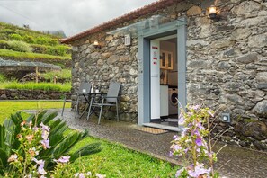 A stone building with a glass door, outdoor seating, and a view of a green hillside. - Casa da Maricas – São Vicente: Tranquility in Madeira’s Natural Paradise (São Vicente)