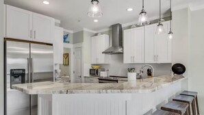 A modern kitchen with white cabinets, a stainless steel refrigerator, a marble countertop, and bar stools. - Beach Pointe 203 (Destin)
