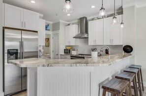 A modern kitchen with white cabinets, a stainless steel refrigerator, a marble countertop, and bar stools. - Beach Pointe 203 (Destin)
