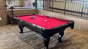 Een pooltafel met een rode viltbekleding, omgeven door een kamerbreed tapijt en een zwarte metalen reling.