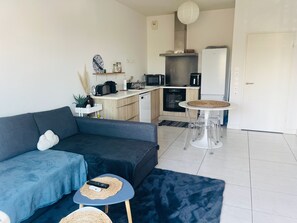 A compact living space with a sofa, a small coffee table, a dining table with chairs, a kitchen area with appliances, and a door. - Anglet Cinq Cantons - T2 with balcony (Anglet)