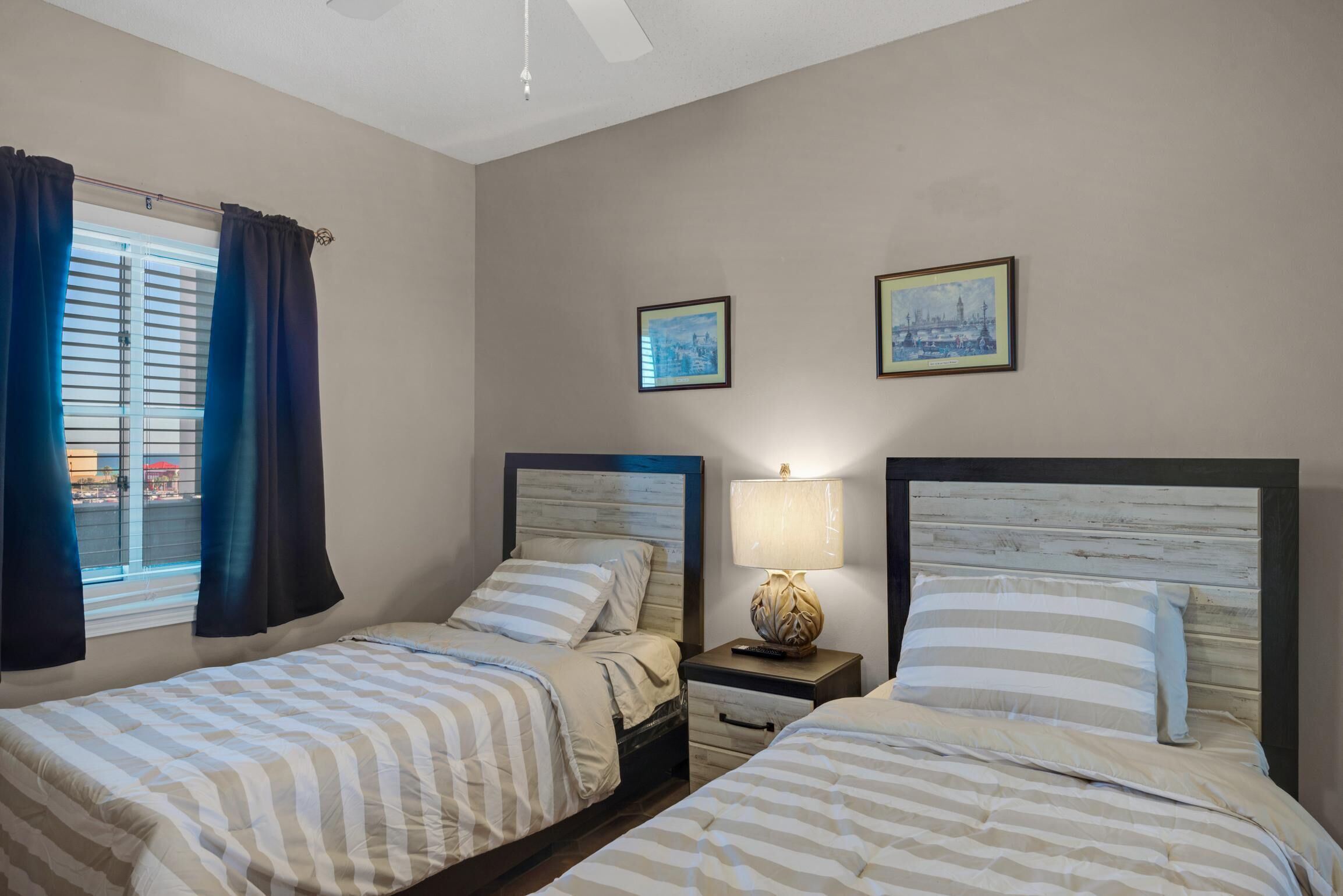 A hotel room with two beds, a window with curtains, a nightstand with a lamp, and two framed pictures on the wall.