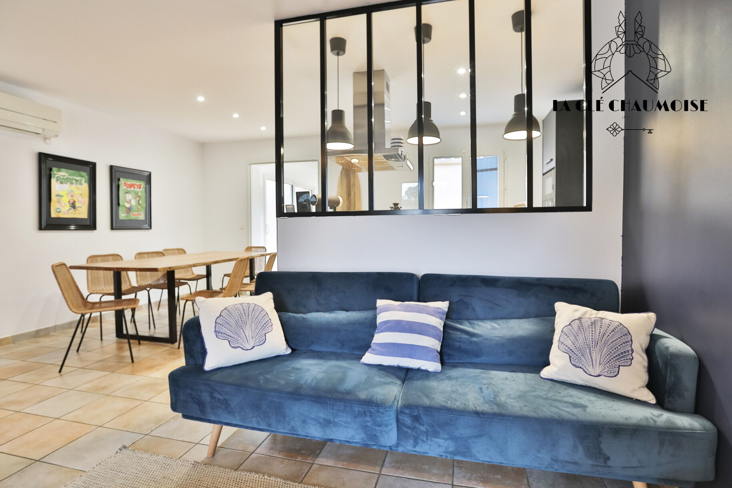 A modern living room with a blue sofa, two framed posters, a dining table with chairs, and pendant lights.