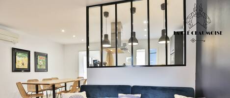 A modern living room with a blue sofa, two framed posters, a dining table with chairs, and pendant lights.
