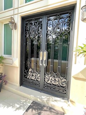 A double metal gate with intricate designs, a potted plant to the left, and a building with a light-colored facade. - Villa Bianca Beachfront Luxury Living w Gym Chef (Cancun)