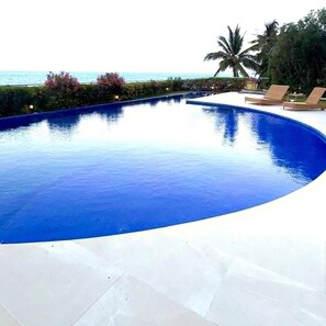 A swimming pool with a curved edge, surrounded by a white deck, palm trees, and a view of the ocean. - Villa Bianca Beachfront Luxury Living w Gym Chef (Cancun)