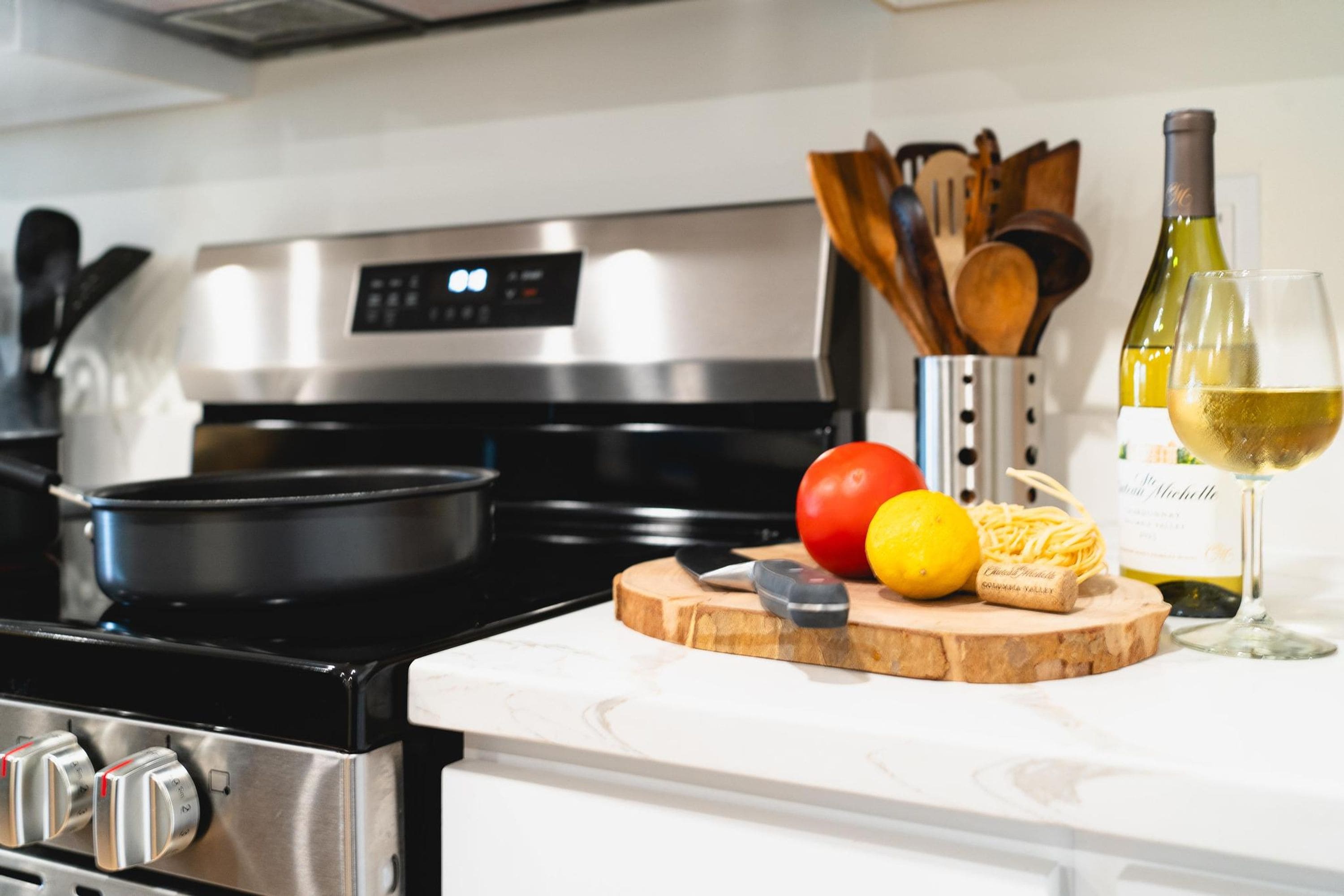 A kitchen with a stove, a cutting board with food items, a wine bottle, and a glass.