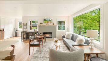A modern living room with a fireplace, a sofa, a dining area, and large windows offering a view of greenery.