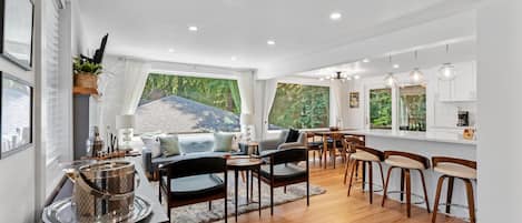 Un amplio espacio de sala con una mesa de comedor, sillas y una zona de cocina con armarios blancos y lámparas colgantes.
