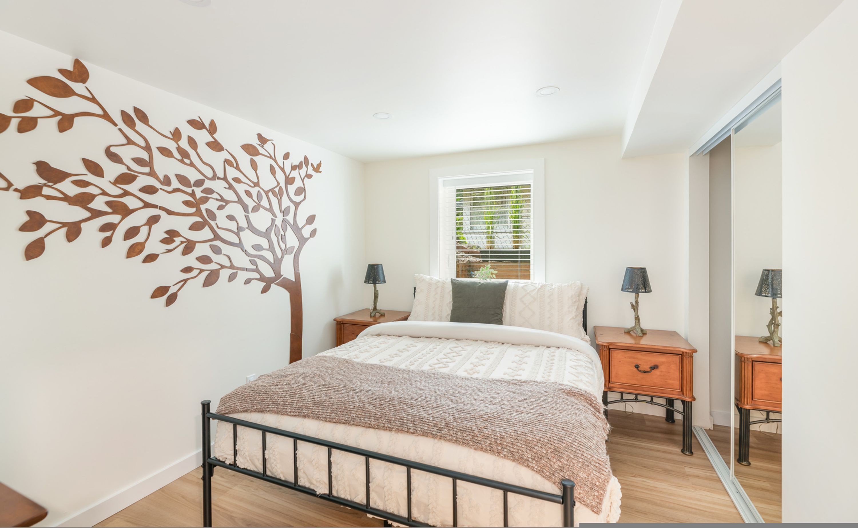 A bedroom with a bed, a wall-mounted tree decoration, a window with blinds, two bedside tables with lamps, and a mirror on the wall.