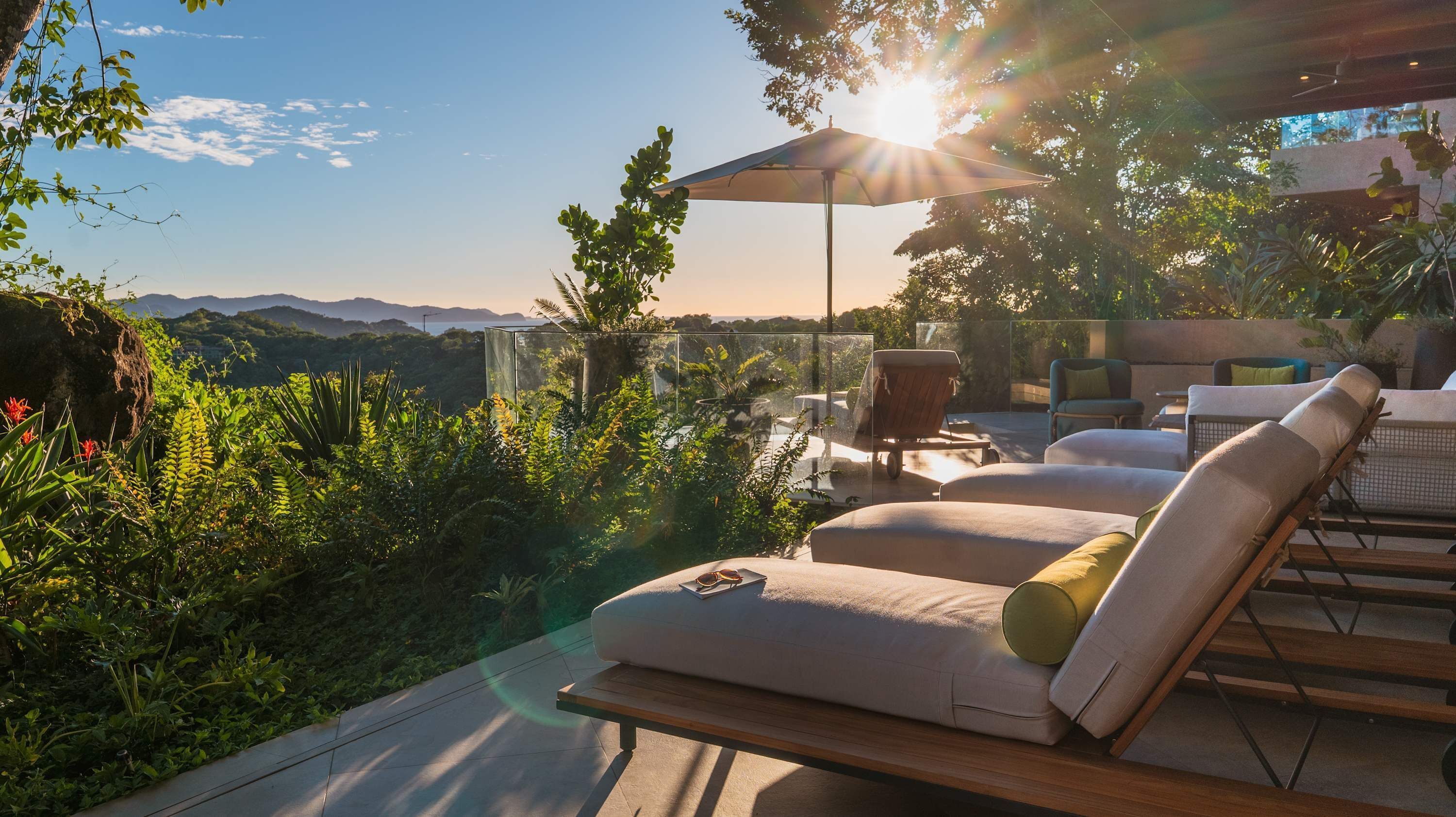A sunlit outdoor seating area with a view of mountains, a glass enclosure, and a wooden table.