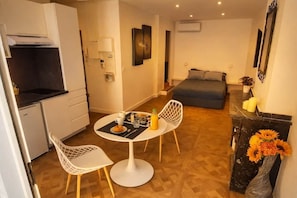Ein kompaktes Hotelzimmer mit Küchenzeile, einem Essbereich mit Tisch und Stühlen, einem Bett und einer kleinen dekorativen Vase mit Blumen.