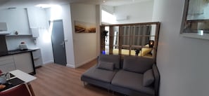 A modern living space with a grey sofa, a small kitchen area, and a large window with curtains. - Studio de la Comédie, top floor (Narbonne)
