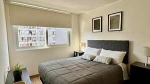 Een slaapkamer met een groot bed, een nachtkastje, een raam met uitzicht op gebouwen en twee ingelijste foto's aan de muur.