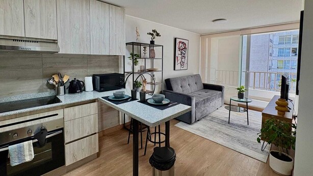 Een moderne keuken met een eethoek, een grijze bank en een raam met uitzicht op gebouwen.
