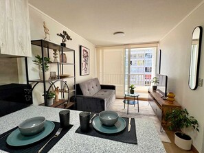 A modern living room with a sofa, a dining table set for two, a bookshelf, and a view of a cityscape through a large window.