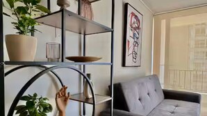 Een woonkamer met een grijze bank, een metalen open kast met planten, en een gekaderd abstract schilderij aan de muur.