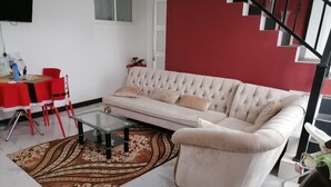 Una sala de estar con un sofá, una mesa de centro de vidrio, una mesa de comedor roja y una escalera.