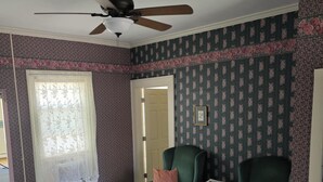 A bedroom with a bed, two green armchairs, a ceiling fan, floral wallpaper, and a window with lace curtains.