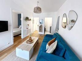 Una sala de estar moderna con un sofá azul, una mesa de centro y un televisor. Hay una puerta que conduce a otra habitación.
