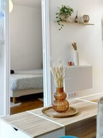 Una habitación moderna con una mesita de noche de madera, un jarrón con plantas secas, una cama con colcha estampada y un aparador blanco con espejo.