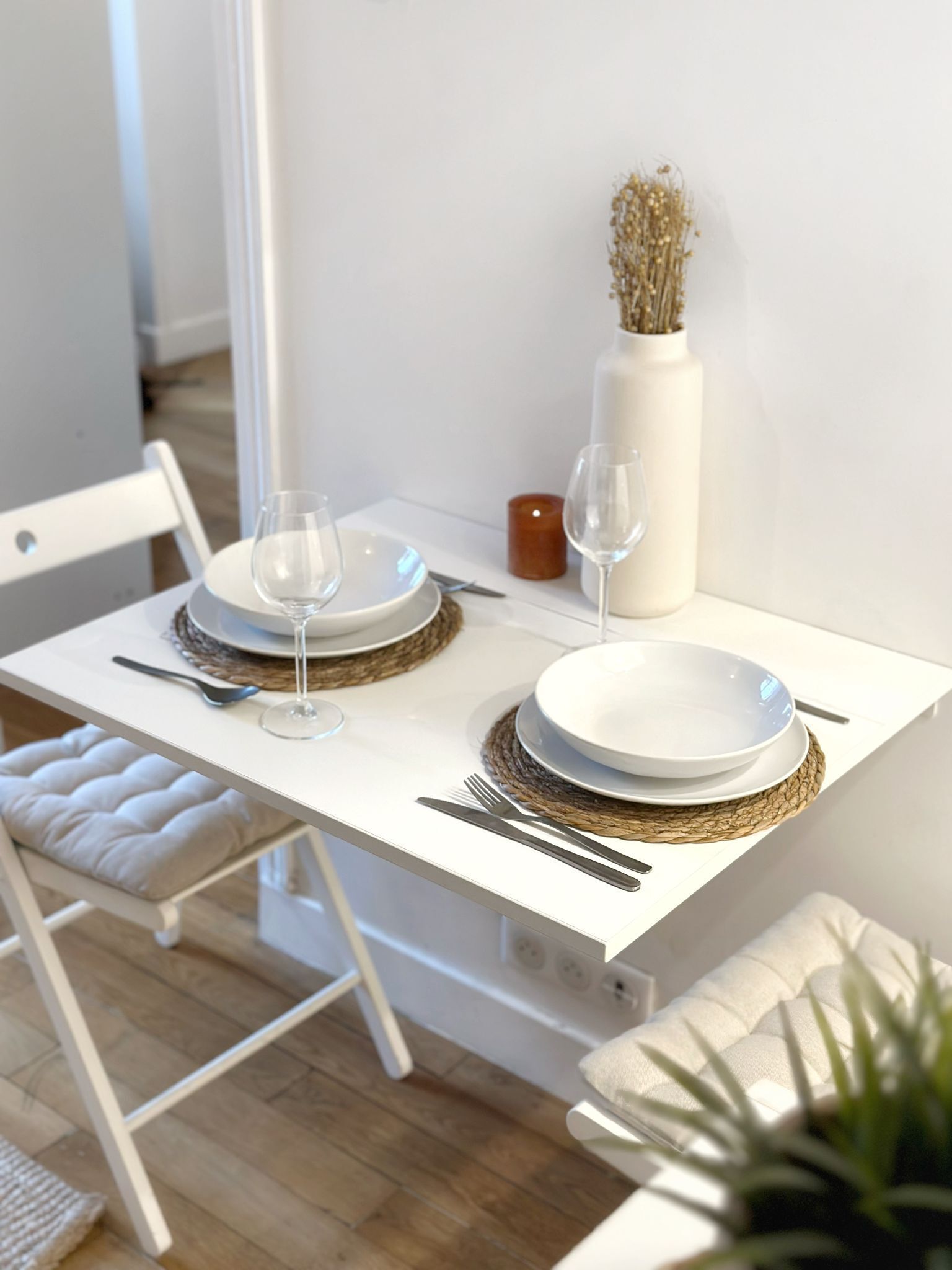 A dining table set with white plates, silverware, and clear wine glasses, accompanied by a vase with dried flowers and a candle.