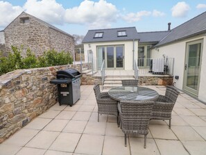 Eine Terrasse mit Steinmauer, Grill, Korbstühlen und einem Glastisch.
