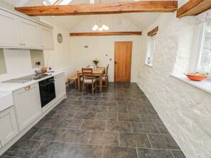 A kitchen with white cabinets, a wooden dining table, and a stone-tiled floor. - Barley Cottage (Swansea)
